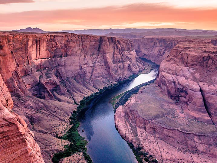 Aerial view of a deep canyon with a winding river at sunset, featuring rugged red rock cliffs and a calm blue stream below